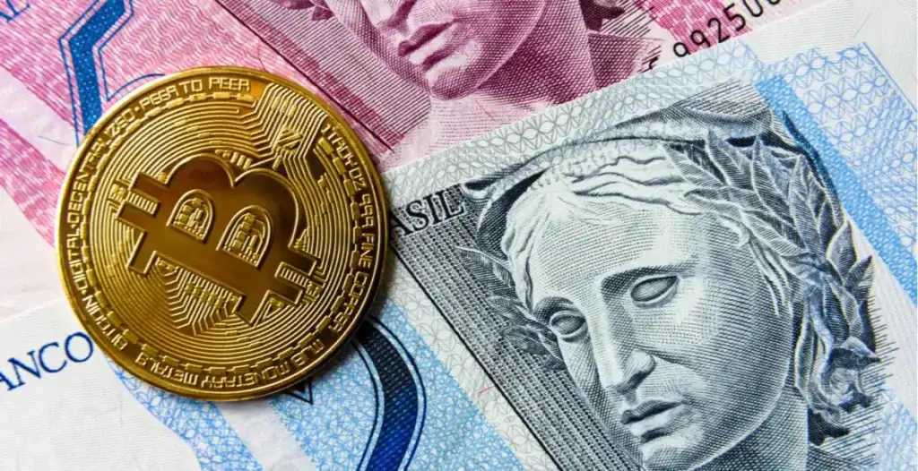 Moeda de Bitcoin posicionada sobre notas de real brasileiras de diferentes valores.